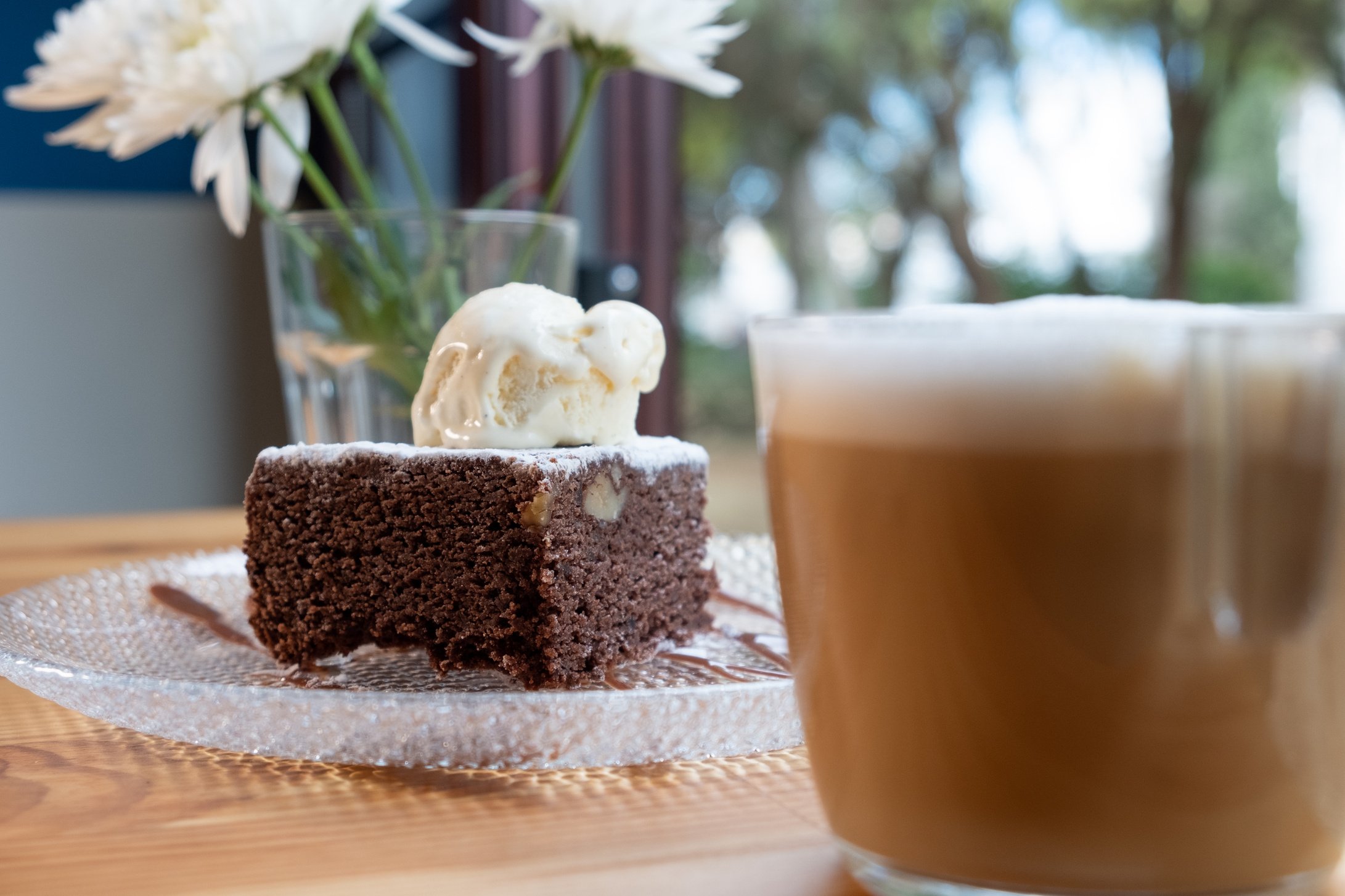 Brownie con helado de vainilla y cafe latte con espuma muy caliente. Postre delicioso vegetariano y natural lleno de proteinas para deportistas. Fondo desenfocado con flores.